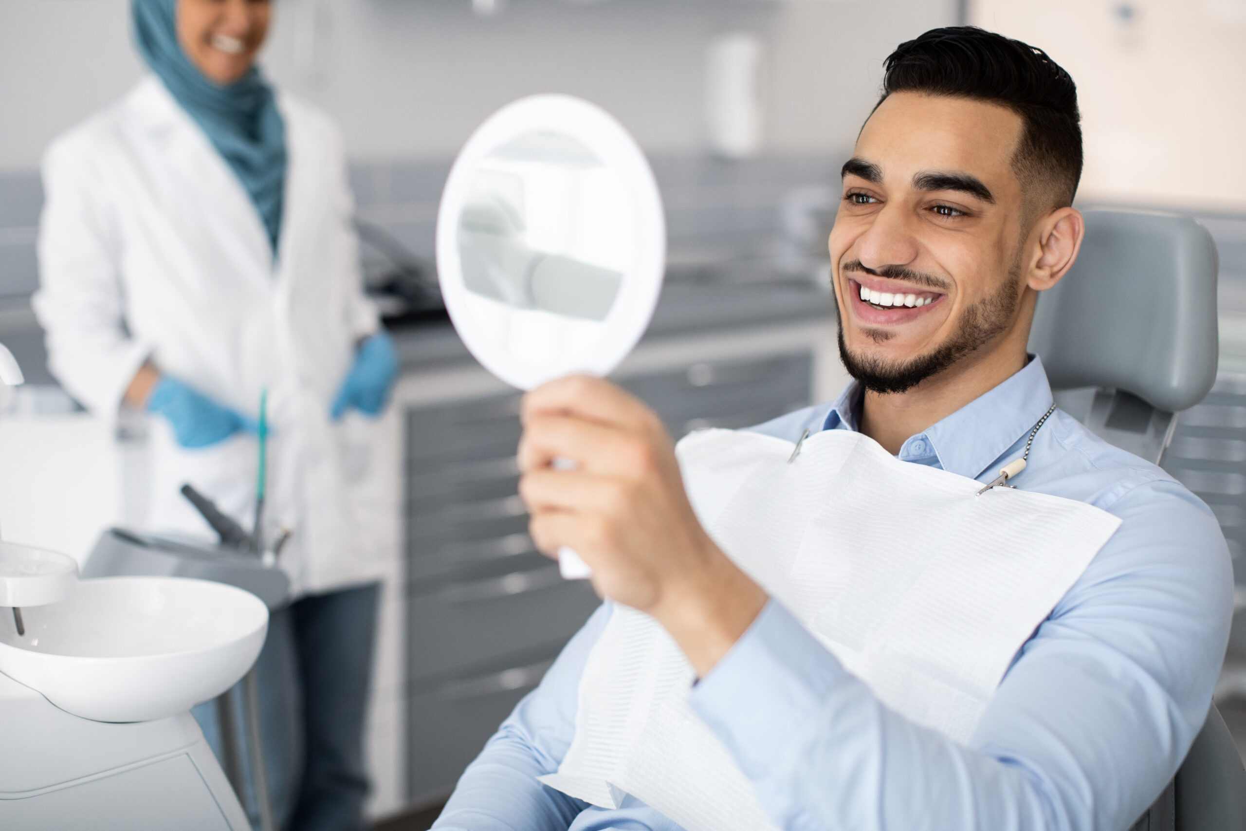 Happy Handsome Arab Man Looking To Mirror After Teeth Treatment In Modern Clinic, Middle Eastern Male Patient Sitting In Chair In Stomatological Cabinet And Enjoying His New Smile, Closeup