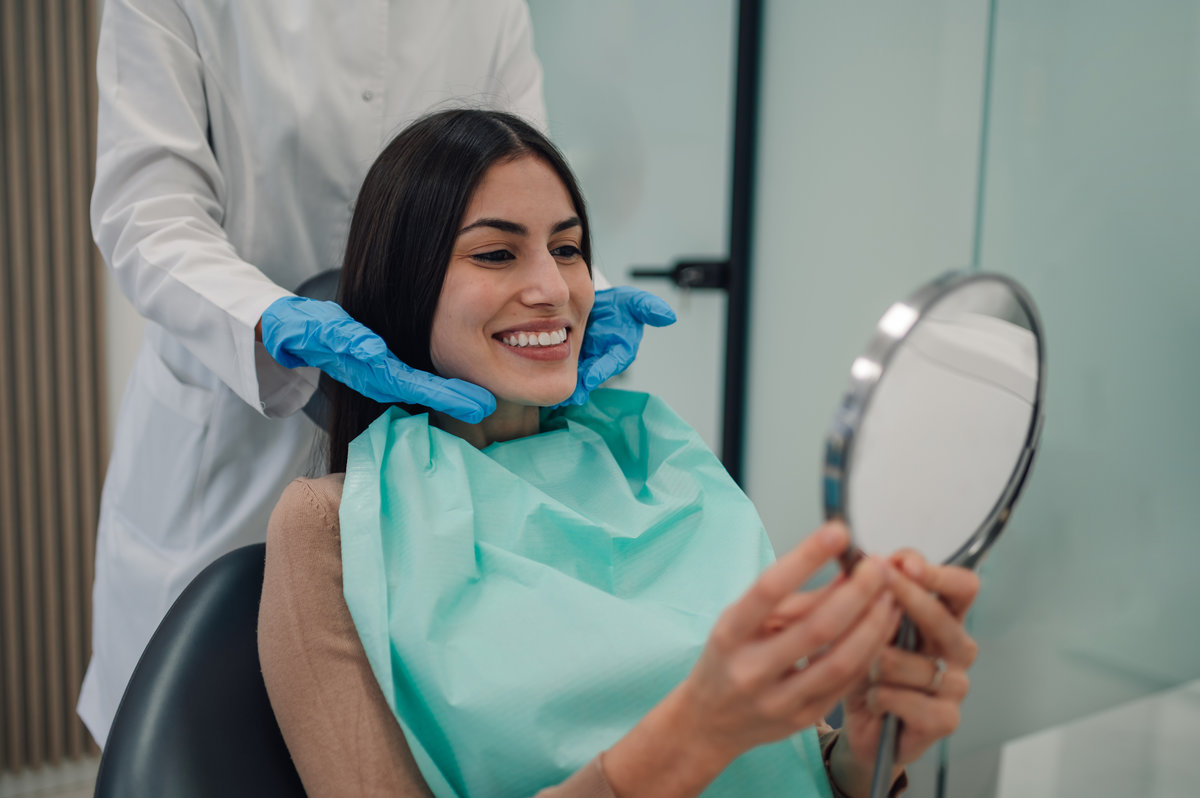 Dentist wearing blue medical gloves gently holding a woman's face as she admires her radiant smile in the mirror after completing a dental procedure at the clinic