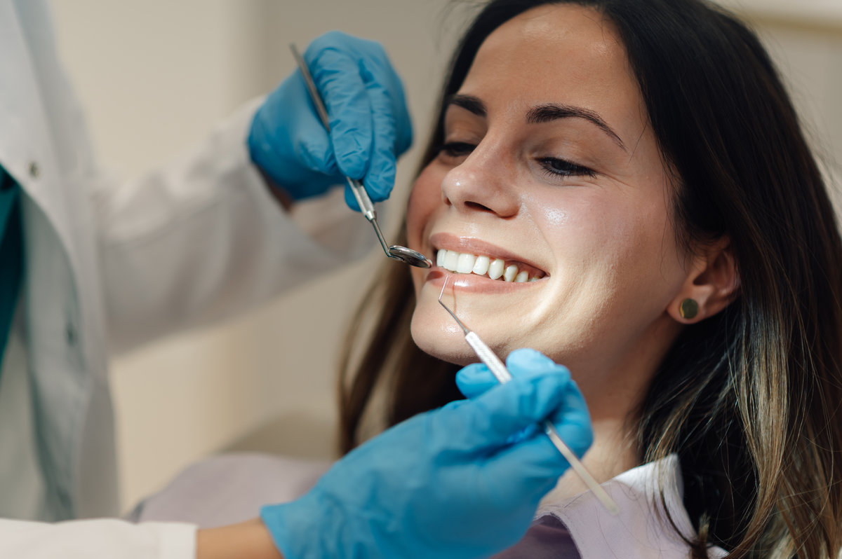 Dentist wearing blue gloves examining teeth of smiling young woman during a thorough dental checkup in a bright, clean clinic, ensuring optimal oral health and hygiene