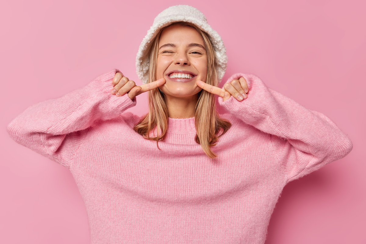 Look at my shining smile. Positive fair haired European woman points at her white teeth smiles broadly wears fashionable panama and casual jumper isolated over pink background has happy mood