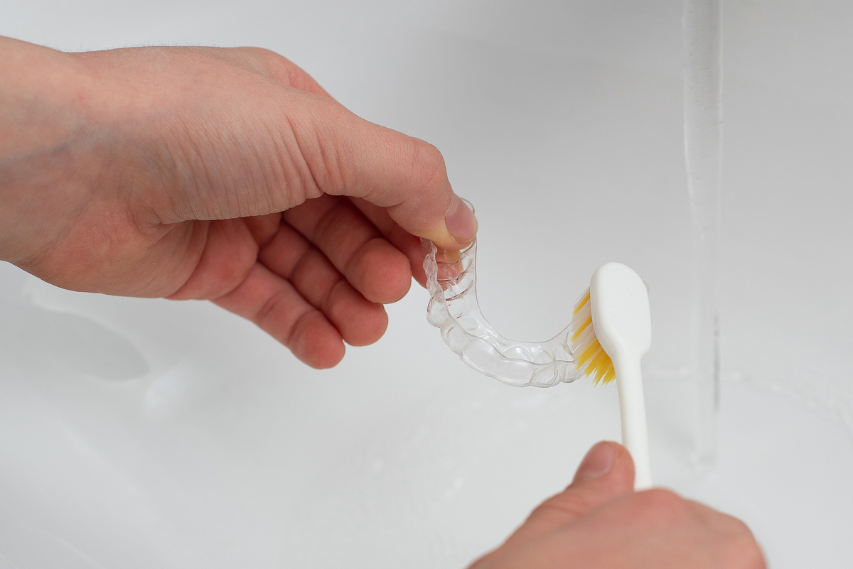 Hands clean aligners with a toothbrush under running water. Home care for aligners