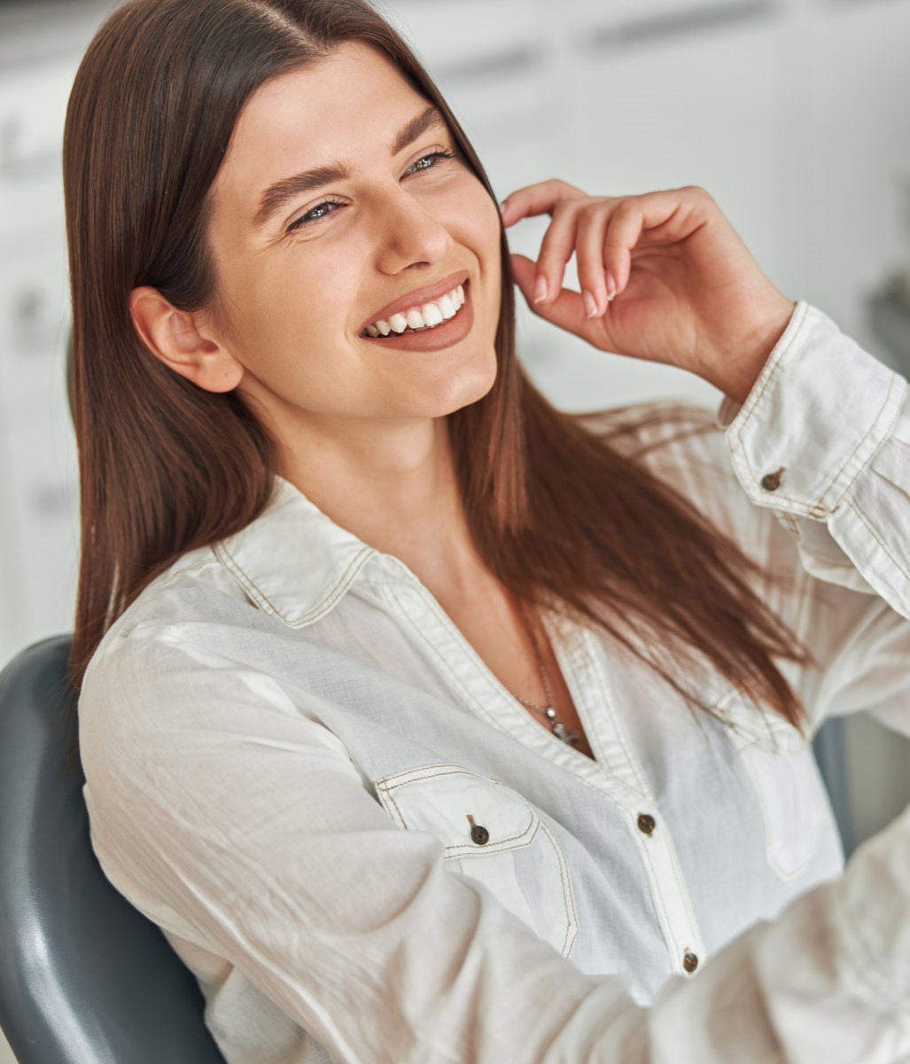 Happy young woman smiling checking out her perfect healthy teeth in the mirror, sitting in a dental chair at the dentist office