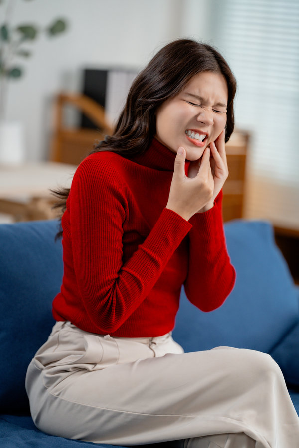 Asian woman having toothache, touching outside mouth with fingers, feeling painful, sitting on sofa at home